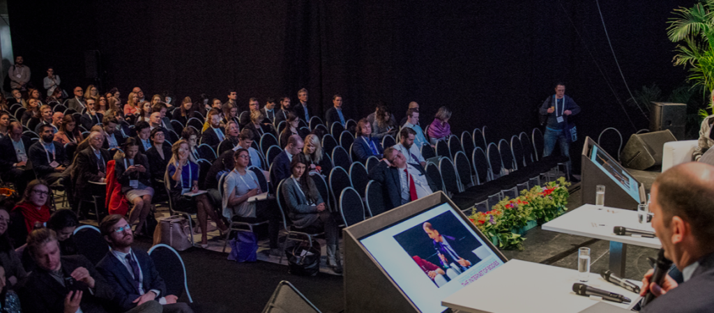 Fotografia do público da conferência, vista a partir do palco de painelistas. Pessoas sentadas em cadeiras assistindo a painelistas